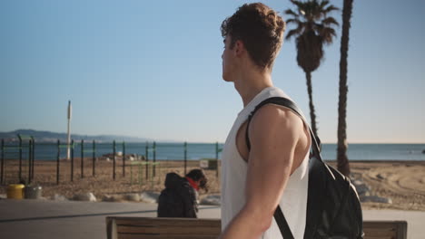 Side-view-shot-of-young-handsome-sporty-man-with-backpack-happily-walking-along-seaside