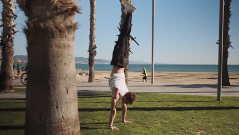 Tracking-around-shot-of-young-handsome-sporty-man-standing-on-hands-during-workout-on-seaside