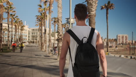 Back-view-shot-of-young-fit-guy-with-backpack-walking-along-seaside