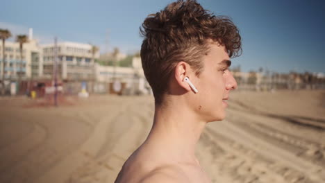 Side-view-shot-of-young-sporty-man-in-wireless-earphones-resting-after-workout-on-beach