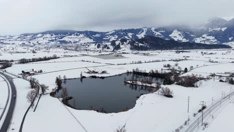 Aerial-orbiting-shot-of-lake-in-Swiss-snowy-winter-landscape-and-alps-in-backdrop