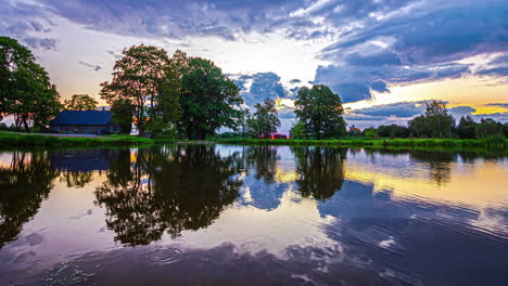 Timelapse-shot-of-white-clouds-passing-by-over-a-lake-beside-wooden-cottage-along-rural-countryside-throughout-the-day