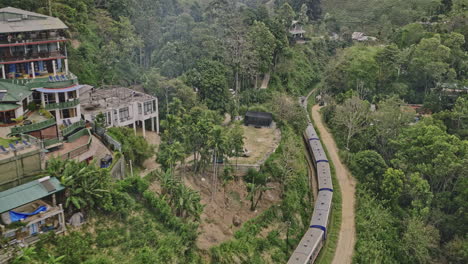 Ella-Sri-Lanka-Aerial-v19-cinematic-birds-eye-view,-drone-following-the-trails-of-train,-passing-through-hillside-resort-hotels,-entering-the-railway-station---Shot-with-Mavic-3-Cine---April-2023