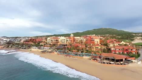 Aerial-view-of-a-luxury-beachfront-resort-in-Cabo,-Mexico,-as-the-waves-roll-in-on-the-sandy-beach,-under-a-beautiful-blue-sky