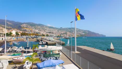 Ondeando-La-Bandera-De-Funchal-En-El-Puerto-Con-Barcos-Atracando-Y-Montañas-En-El-Fondo-Durante-El-Día-Soleado---Portugal,-Europa