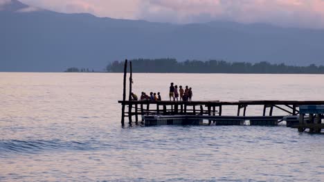 Niños-Viendo-La-Hermosa-Puesta-De-Sol-En-El-Muelle-Con-Vistas-Al-Agua-Del-Océano-En-Una-Remota-Isla-Tropical-En-Raja-Ampat,-Papúa-Occidental-En-Indonesia