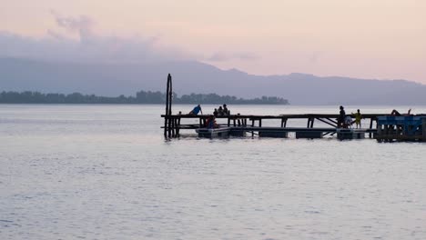 Grupo-De-Personas-Relajándose-En-El-Muelle-Con-Vista-Al-Océano-Durante-La-Hermosa-Puesta-De-Sol-Con-Cielo-Rosado-En-La-Isla-Tropical-En-Raja-Ampat,-Papua-Occidental,-Indonesia