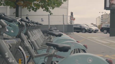 Bicicletas-Eléctricas-Velib-Estacionadas-En-Línea,-Autos-Pasando-Al-Fondo