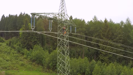 Trabajador-Subiendo-Una-Torre-De-Energía-De-Alto-Voltaje-En-Medio-De-Un-Bosque-Verde,-Día-Nublado,-Toma-Aérea