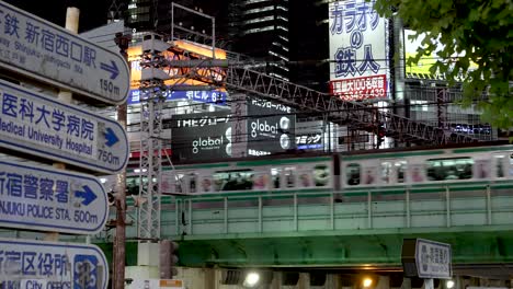 Yamanote-Line-Train-Passing-On-Elevated-Train-Track-In-Shinjuku-At-Night-With-View-Of-Information-Road-Signs-In-View