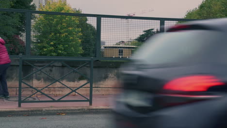 Ältere-Frau-Schiebt-Einen-Trolley-Durch-Die-Straße,-Im-Vordergrund-Vorbeifahrende-Menschen-Und-Autos