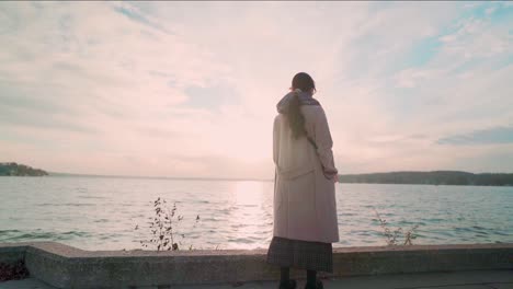 Standing-Asian-woman-in-coat-turn-around-and-look-at-the-sunset-at-lake-in-fall-evening-slow-dolly-out