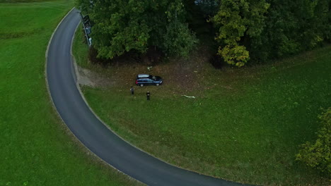 Aerial-unidentified-people-near-blue-car-in-grass-field-near-curbed-road-and-forest