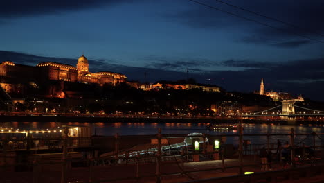 Ein-Wunderschöner-Abendblick-Auf-Buda-Mit-Dem-In-Orangefarbenem-Licht-Beleuchteten-Burgberg,-Während-Flussschiffe-Auf-Der-Donau-Fahren