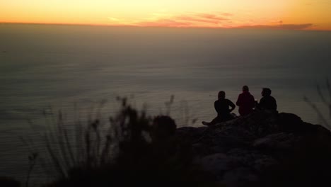 Die-Silhouette-Dreier-Junger-Freunde-Trinkt-Einen-Sundowner-Auf-Einem-Berggipfel-Während-Eines-Wunderschönen-Orangefarbenen-Sonnenuntergangs-Mit-Rollenden-Wolken