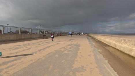 Corredores-De-Caridad-Corriendo-A-Lo-Largo-Del-Paseo-Marítimo-Junto-A-Una-Playa-De-Arena-Con-Nubes-De-Tormenta-Grises-Y-Un-Sol-Brillante