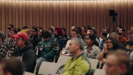 Eine-Große-Gruppe-Von-Tagungsteilnehmern,-Publikum,-Menschenjacken,-Die-In-Einem-Präsentationszentrum-Auf-Einen-Konferenzredner-Warten,-Mit-Einer-Kamera-Im-Hintergrund,-Einer-Wunderschön-Beleuchteten-Universität