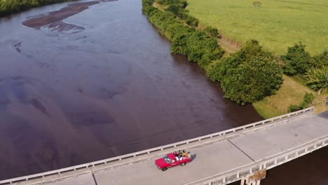 Pickup-truck-full-of-people-riding-in-back-passes-tuk-tuk-while-crossing-bridge-over-river