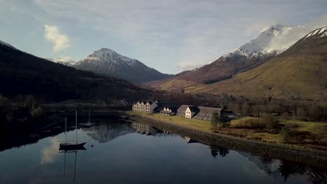 Aerial-approaching-shot-of-Isles-of-Glencoe-Hotel-at-lake-loch-leven-in-scotland-at-sunset-time---Snowy-mountains-in-background---descending-flight