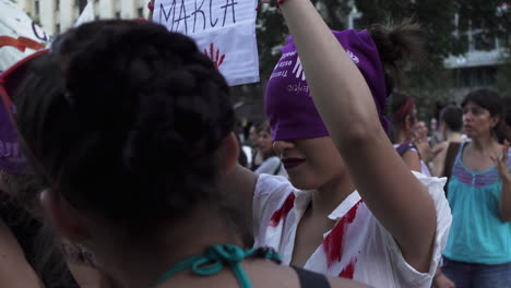 Eine-Junge-Frau-Führt-Eine-öffentliche-Kundgebung-Durch,-Hält-Ein-Plakat-In-Der-Hand,-Ihre-Augen-Sind-Mit-Einem-Lila-Feministischen-Kopftuch-Bedeckt,-Sie-Hat-Rote-Flecken,-Die-Blut-Und-Gewalt-Darstellen