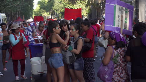 Una-Joven-Pinta-La-Cara-De-Una-Amiga-Con-Brillo-Verde-En-Una-Marcha-Pública-Por-Los-Derechos-De-Las-Mujeres-En-Cámara-Lenta.