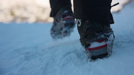 Low-angle-hiker-with-ice-cleats-and-camera-gimbal-walks-away-toward-snowy-peak