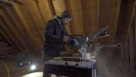 Hombre-Con-Gorro-Y-Chaqueta-Usando-Una-Sierra-Ingletadora-En-Un-Taller-De-Madera,-Polvo-En-El-Aire,-Iluminación-Interior