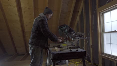Man-in-beanie-and-plaid-shirt-using-a-chop-saw-in-a-woodshop-with-window-light