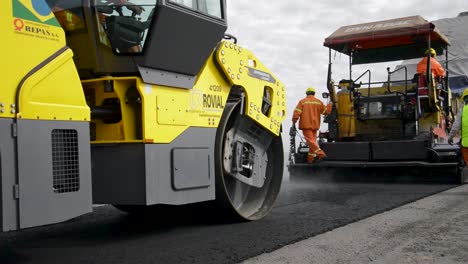 Trabajadores-De-La-Construcción-Con-Ropa-De-Alta-Visibilidad-Operando-Maquinaria-De-Pavimentación-En-Una-Carretera-Nueva,-Día-Nublado-Cerca-De-Buenos-Aires