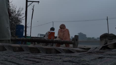 Rear-outdoor-view-of-old-man-and-a-woman-sitting-on-a-bed-made-of-jute-ropes-during-sunset-beside-streets-of-Chichawatni-city-in-Punjab,-Pakistan