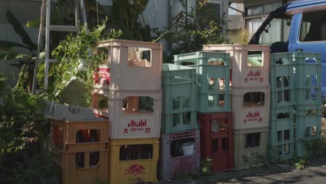 Stacked-beverage-crates-in-a-back-alley-with-foliage-in-Izumi,-Kyushu,-Japan