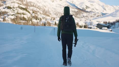 Man-in-winter-jacket-with-backpack-and-gimbal-walks-through-snow-toward-buildings