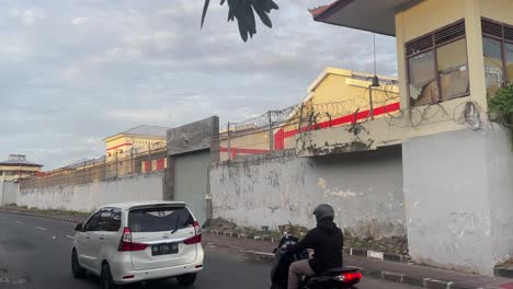 Panning-shot-of-a-Jail-as-trafic-goes-by-in-front-of-tall-concrete-walls-with-barbed-wire