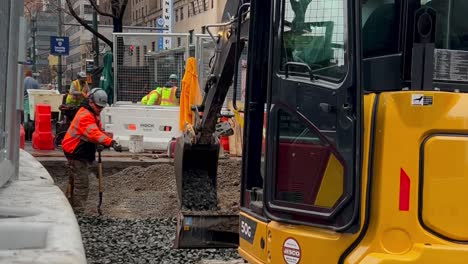 Eine-Aufnahme-Aus-Einem-Niedrigen-Winkel-Zeigt-Einen-Mann,-Der-Auf-Einer-Baustelle-In-Manhattan,-New-York,-Kies-Schaufelt,-Während-Ihm-Ein-Anderer-Mann-In-Einem-Bagger-Hilft