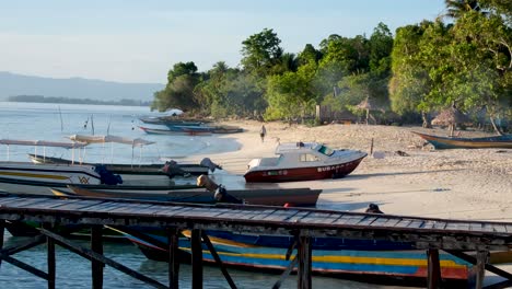 Vista-Panorámica-Del-Paisaje-De-Una-Sola-Persona-Caminando-Por-Una-Playa-De-Arena-Blanca-Y-Barcos-De-Pesca-Amarrados-En-Una-Isla-Tropical-Cubierta-De-árboles-De-Selva-Tropical.