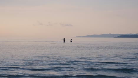 Small-group-of-women-with-umbrella-for-sun-protection-walking-across-ocean-lagoon-between-two-tropical-islands-at-low-tide