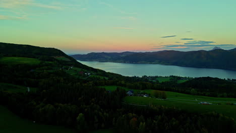 Aerial-shot-of-a-lake-surrounded-by-mountains-and-some-small-houses-in-the-colorful-sunset-sky