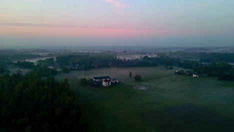 Panoramic-aerial-shot-of-a-huge-green-field-with-some-houses,-surrounded-by-nature,-trees-and-fog