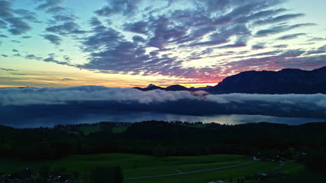 Drone-shot-of-the-colorful-sky-with-clouds-over-a-natural-field-with-trees-and-a-lake-surrounded-by-mountains