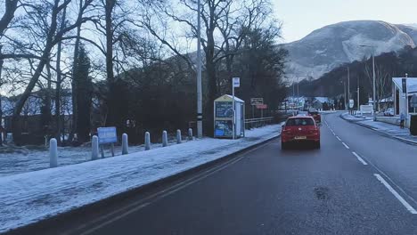 Winter-day-in-Skye-with-vehicles-on-snow-lined-road,-mountains-in-background,-early-morning-light,-car-POV