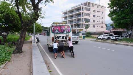 Group-of-Timorese-young-men-pushing-a-broken-down-district-bus-on-the-streets-with-passing-traffic-in-capital-city-of-East-Timor,-Southeast-Asia