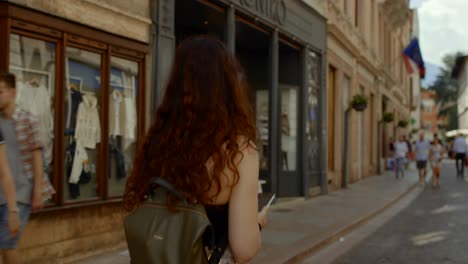 Toma-De-Seguimiento-De-Una-Mujer-Joven-Con-Cabello-Ondulado-Caminando-Por-Una-Calle-Con-Tiendas-En-Verona,-Italia.