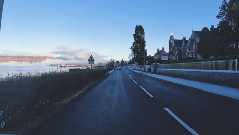 Frosty-morning-on-Skye-with-cars-on-road,-historical-buildings-and-tranquil-sea-under-blue-skies,-car-POV
