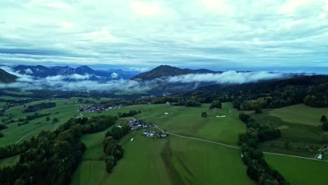 Hermosa-Toma-Aérea-De-Un-Enorme-Campo-Verde-Con-Algunas-Casas,-Rodeada-De-Montañas,-Naturaleza-Y-Nubes-Bajas