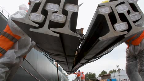 Pair-of-workers-in-safety-suits-and-hardhats-move-steel-beams-off-crane-of-boat-in-port