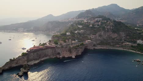 Aerial-shot-over-the-ocean-while-a-drone-orbiting-around-an-island-at-sunset-near-Taormina,-Sicily