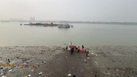 Vista-Trasera-De-Una-Toma-Amplia-De-Personas-Bañándose-Y-Ofreciendo-Oraciones-A-Dios-Cerca-Del-Río-Hooghly-Con-Paisaje-Urbano-Al-Fondo-En-Kolkata,-India.