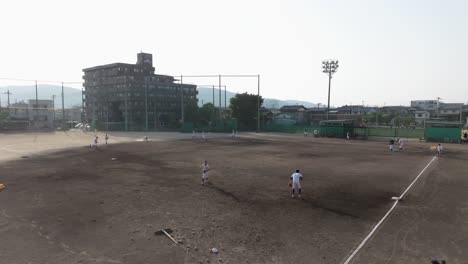 El-Equipo-De-Béisbol-De-La-Escuela-Entrena-Jugando-Partidos-En-El-Campo-De-Tierra,-Vista-Aérea-En-Cámara-Lenta,-Horizonte,-Montañas-Y-Fondo-De-La-Ciudad.