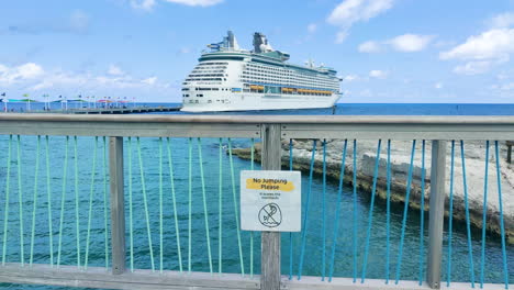 Amidst-the-grandeur-of-docked-cruise-ship,-whimsical-caution-sign-on-the-bridge-adds-touch-of-humor-to-the-bustling-waterfront-scene-|-funny-caution-sign-on-island-with-cruise-ship-docked