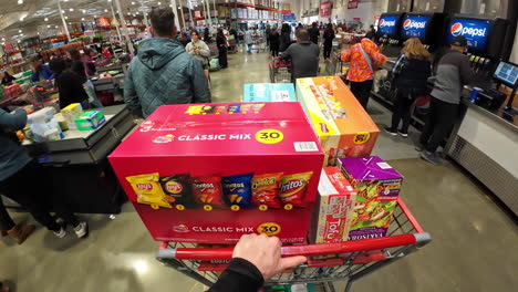 First-Person-shopper-carry-supermarket-cart-full-of-boxes-of-snack-and-groceries-hypermarket-with-customers-and-cashiers